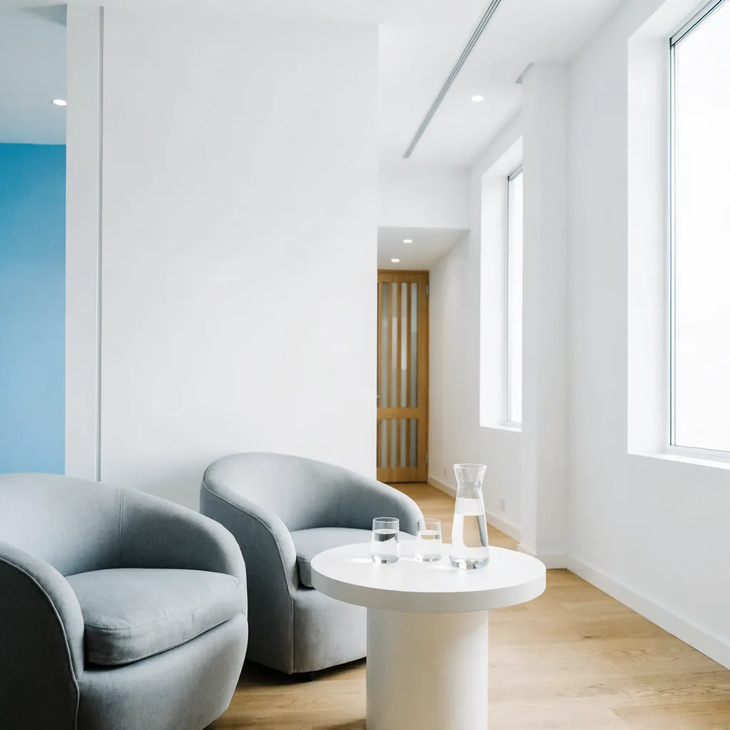 Minimal waiting lounge with two light grey armchairs, a round white table with a water carafe and glasses, pale-blue accen…